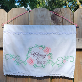 A white fabric wall hanging with pink and green cross stitch floral details and the text 'Whimsy & RAGE' in the center, hanging from a pink cord against a wooden fence