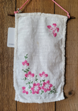 A rectangular white linen cloth with fringed edges, decorated with pink floral embroidery and green stems, hanging on a wooden surface with a pink cord and a small white tag.
