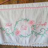 A white cotton wall hanging with embroidered pink script text reading 'What The Fuck?' and a green floral vine border, finished with a pink zigzag trim at the top and a white lace hem, laid flat on a wooden surface.