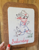 A wooden-framed embroidered art piece featuring a whimsical cowboy dog holding a bone illustration with a red bandana and the word 'Saturday' in red script.