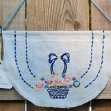 A white linen half-circle wall hanging with blue embroidered borders and a central basket of multicolored flowers, hanging on a wooden fence.