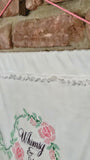 A close-up of a white embroidered fabric panel featuring pink flowers, green leaves, and the cursive text 'Whimsy & Rage' in grey, hung against a rustic brick wall with a pink cord.