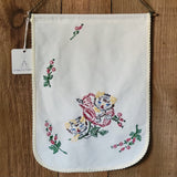 A white cotton wall hanging with scalloped edges and colorful floral embroidery, hanging on a wooden surface