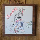 A square framed piece of embroidered art featuring a cartoon dog dressed in cowboy clothing with the word 'Thursday' in red script. The embroidery is on a white background and is housed in a dark brown wood frame, set against a rustic wooden wall.