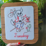 A square wooden plaque featuring a western-themed embroidered dog riding a stick horse, with the word 'Tuesday' stitched in red at the bottom. The background shows green plants and a wooden floor.