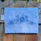 A square fabric wall hanging in light blue with a blue floral pattern, hanging on a wooden fence.