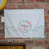 A white embroidered wall hanging with lace trim hangs on a pink cord against a red brick wall. The center features a floral wreath design with pink roses, green leaves, and the words 'Whimsy & RAGE' stitched in dark script.