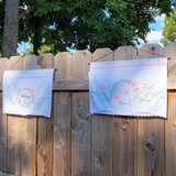 Two white embroidered wall hangings on a wooden fence outdoors. The left flag reads 'whimsy and rage' with pink roses and green vines, and the right one reads 'WTF' with similar floral embroidery
