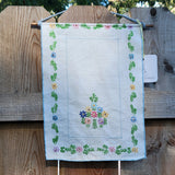 A square white fabric wall hanging with embroidered floral borders and a central flower motif, displayed against a wooden fence.