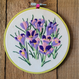 A circular embroidery hoop holding a floral embroidery in purple, pink, and green on cream fabric, photographed against a rustic wooden background.