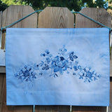 A rectangular light blue fabric banner with a blue floral vine print, hanging from a rope against a wooden fence outdoors.