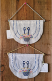 Two handmade fabric wall hangings displayed on a wooden wall. Each white fabric panel is shaped like a semicircle with a hanging cord, a wooden dowel, and an embroidered design of a blue basket with flowers and a bow.