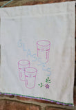 A white fabric tea towel wall hanging with embroidered designs of three glasses, the word 'GLASSES' in blue, and small purple and green flowers, hanging on a brick wall