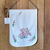 A white fabric wall hanging with embroidered floral motifs and a scalloped edge, suspended by twine against a wooden plank background. 