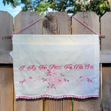 An off-white embroidered wall hanging piece with pink text reading 'I Am Too Pretty For This Shit' and pink floral embroidery, suspended by pink strings against a wooden fence outdoors.