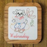 A square wooden plaque featuring a hand-stitched cartoon dog dressed in western clothing, holding a cowboy hat and rope, with the word 'Wednesday' stitched in red at the bottom. The plaque is placed on a dark wood surface.