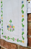 A white embroidered cloth napkin or handkerchief with a colorful floral and leaf border, hanging against a rustic brick wall.