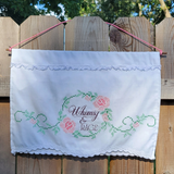 A white fabric wall hanging on a pink cord against a wooden fence. The flag features embroidered pink roses, green leaves, and the words 'Whimsy & RAGE' in a cursive font.