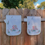 Two white fabric flags with colorful embroidered flowers and leaves, hanging from a wooden fence outdoors.