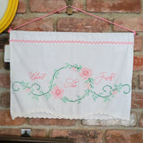 A white fabric wall hanging with pink and green cross stitch floral vines and the hand embroidered words “What The Fuck?” in pink script, suspended by a pink string against a red brick wall